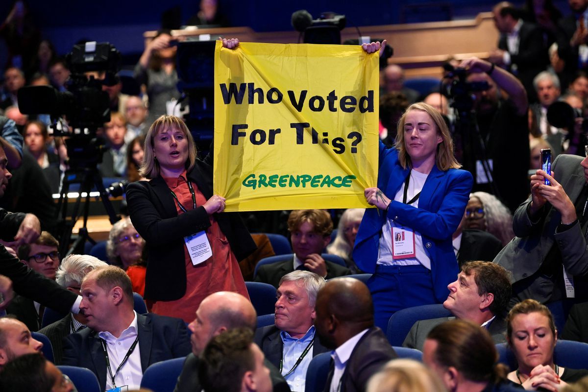 Two white women among audience of people hold up a yellow banner which read "Who Voted for this?" with Greenpeace logo