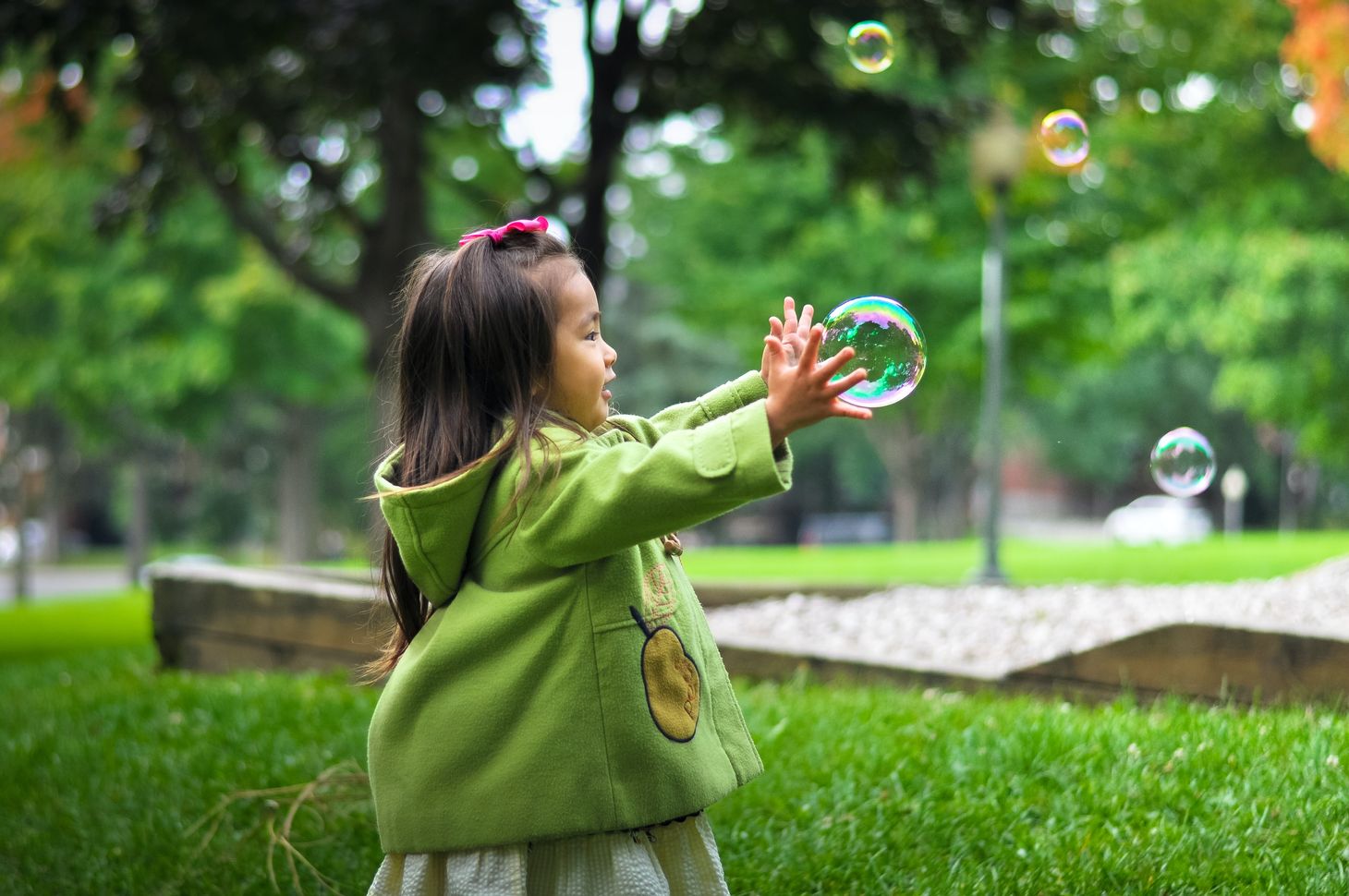 little girl with long dark hair in a pink ribbon wearing a green jacket catching a bubble outside in the park