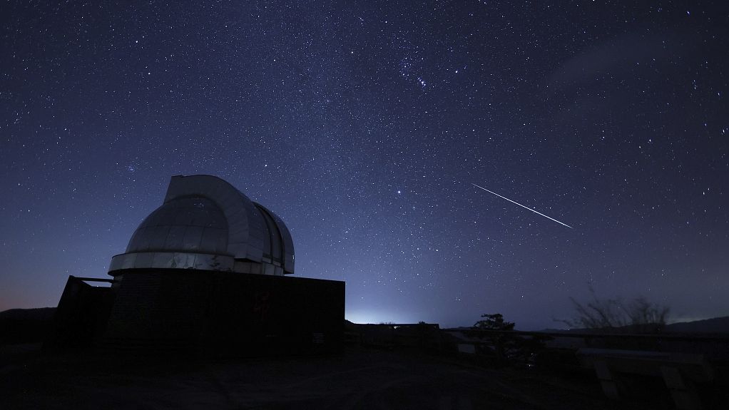 Night sky with outline of an curved building in the foreground. A shooting star streaks from top left to bottom right