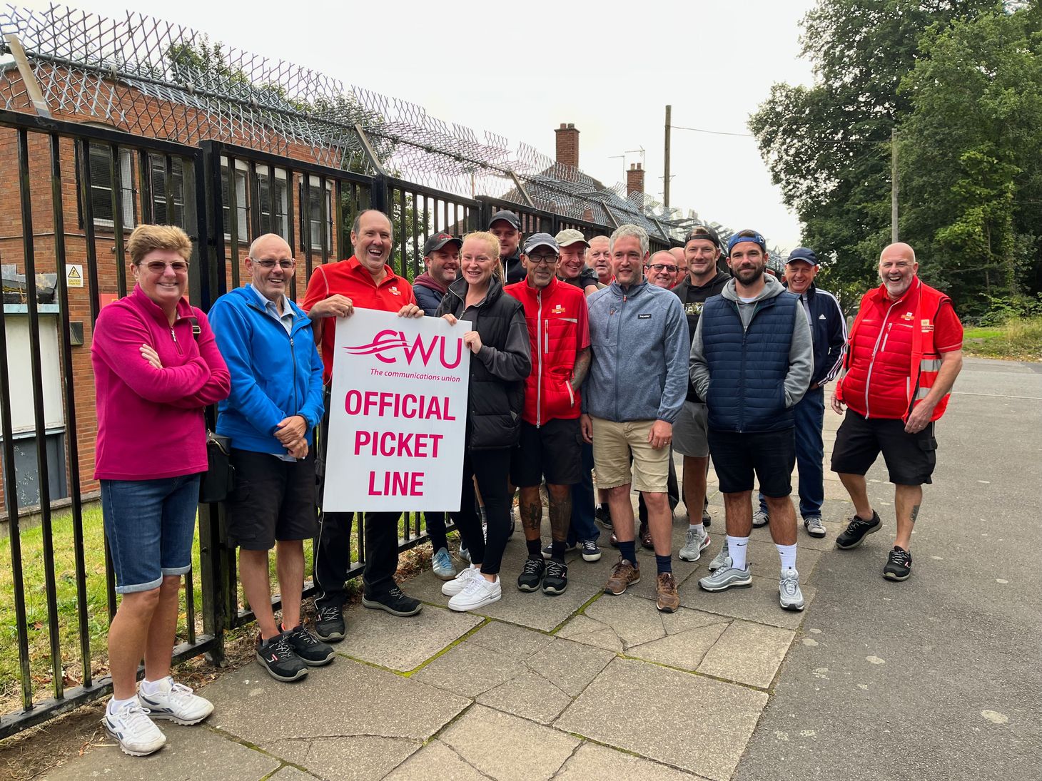 A group of men and women postal workers are standing outside a fence holding a banner which reads CWU Official picket line